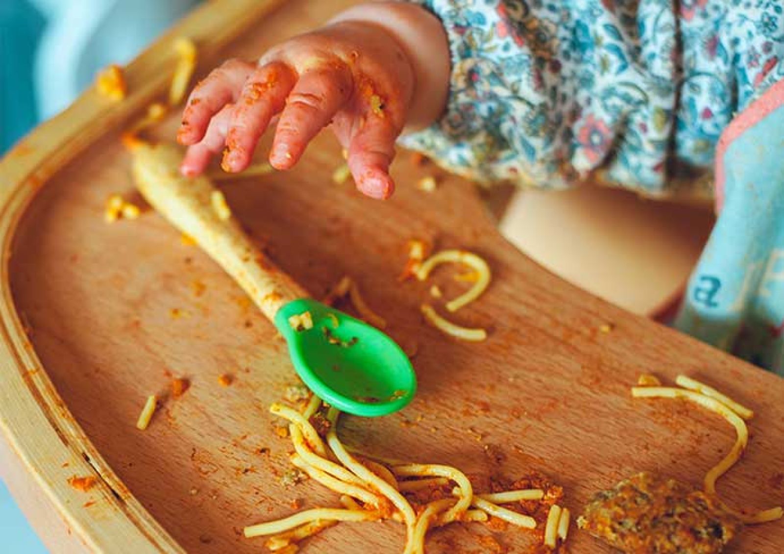 child's hand reaching for a spoon on a plate that is dirty with spaghetti